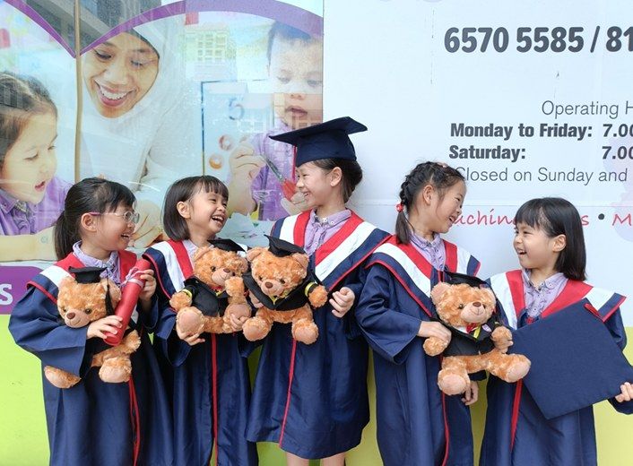 A group of graduating children in caps and gowns smiling together with their teddy bears